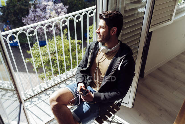 Молодий усміхнений красивий випадковий чоловік в гарнітурі, що спирається на терасові перила зі смартфоном і дивиться вбік — стокове фото