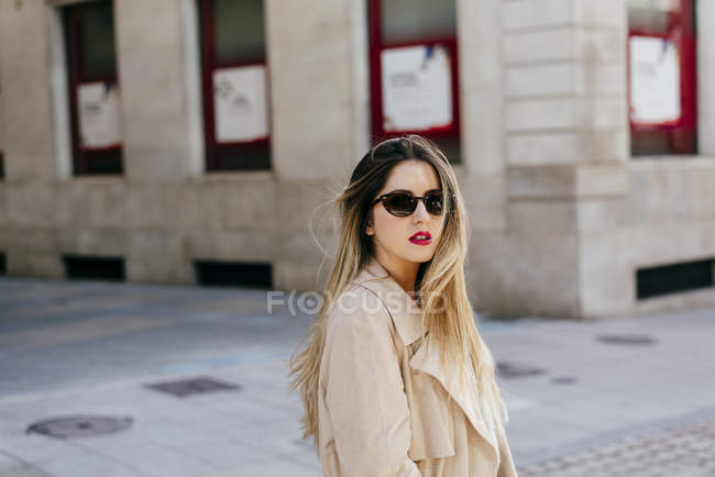Junge hübsche Frau in stylischem Mantel und Sonnenbrille posiert auf der Straße vor einem Marmorgebäude mit leuchtend roten Fenstern — Stockfoto