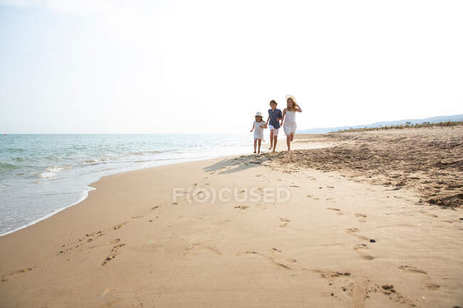 Счастливые и улыбающиеся дети в повседневной одежде бегают босиком по берегу моря на песчаном пляже в летний солнечный день — стоковое фото