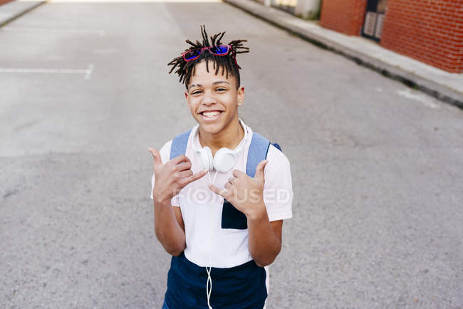 Joven hombre guapo afroamericano alegre en ropa casual con auriculares de pie en la calle y mostrando el signo de la mano shaka - foto de stock