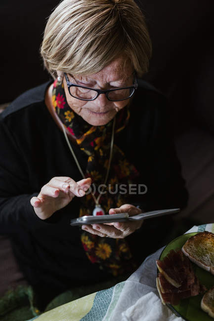 Seniorin schaut und berührt Smartphone-Bildschirm, während sie auf Sofa im Wohnzimmer sitzt — Stockfoto