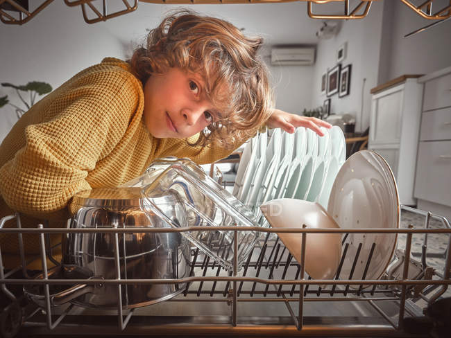 Niño sonriendo mirando dentro de lavavajillas abierto en la cocina - foto de stock