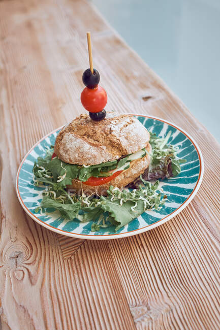 Delizioso panino vegetariano con pomodori, cipolla, lattuga, olive e pomodorini su piatto luminoso su tavolo in legno nel ristorante — Foto stock