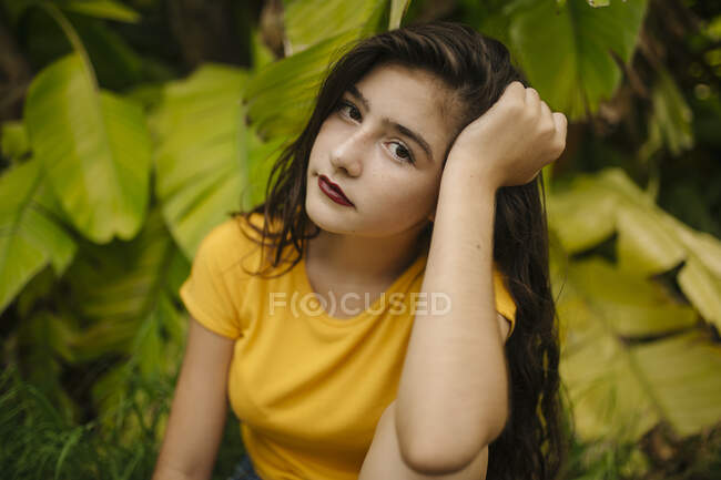 Nachdenkliche junge Frau in gelbem T-Shirt stützt Kopf und blickt in die Kamera, während sie neben exotischem Strauch im Garten sitzt — Stockfoto