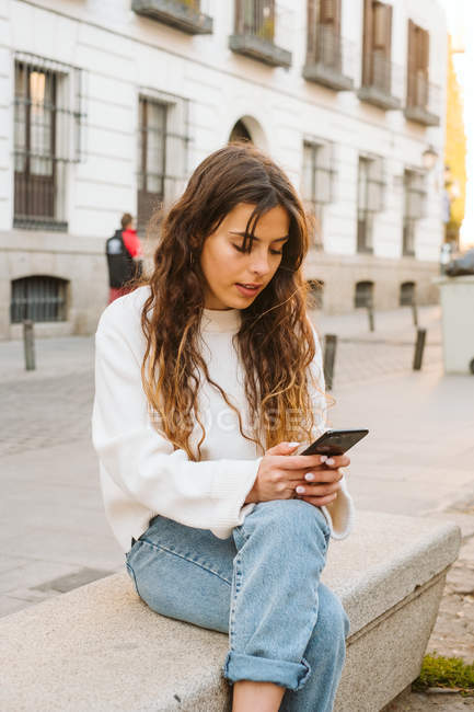 Jovem do sexo feminino em roupa casual sentado e navegando smartphone na rua da cidade — Fotografia de Stock