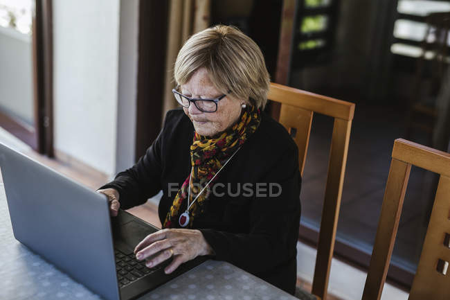 Літня жінка в окулярах переглядає ноутбук, сидячи за столом у затишному будинку — стокове фото