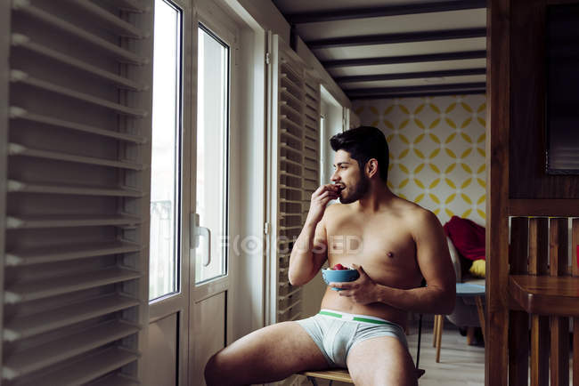Вид сбоку молодого жизнерадостного сексапильного успешного мужчины в нижнем белье, сидящего на деревянном стуле и поедающего клубнику возле балкона — стоковое фото