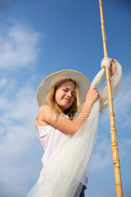 Girl in hat attaching awning on pole with clear blue sky on sand on beach — Stock Photo