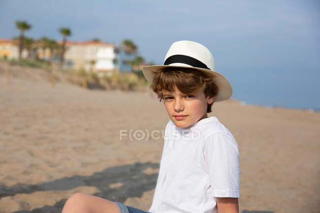 Netter Junge in weißem T-Shirt und Hut sitzt im Sommer am Sandstrand — Stockfoto