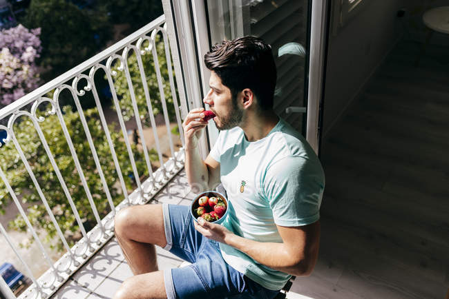 Вид збоку молодого веселого сексуального успішного чоловіка в нижній білизні, що сидить на дерев'яному стільці і їсть полуницю біля балкона — стокове фото
