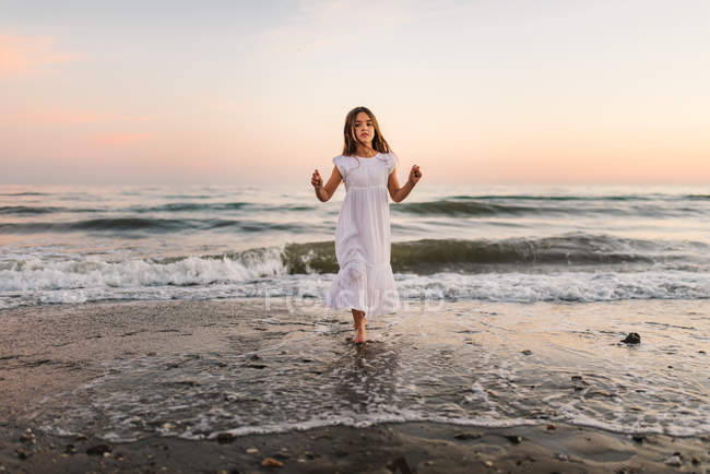 Little girl in white dress walking in water on beach at sunset — Stock Photo