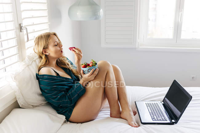 Young happy woman holding strawberry bowl on bed — Stock Photo