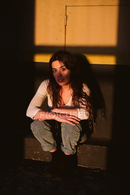 Jeune femme en tenue décontractée assise sur le mur du bâtiment à la lumière du soleil — Photo de stock