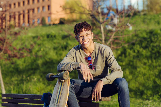 Молодий чорний чоловік сидить на лавці зі скейтбордом — стокове фото