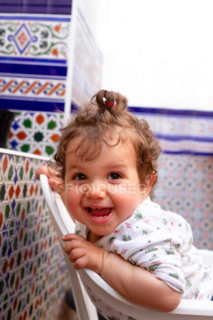 Vista lateral del adorable bebé sonriendo y mirando hacia otro lado mientras se divierten en la silla cerca de la pared ornamental de la terraza - foto de stock