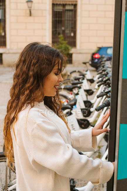 Jeune femme souriante en tenue décontractée en utilisant kiosque sur la station de partage de vélos sur la rue de la ville — Photo de stock