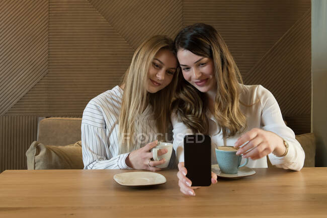 Dos amigas elegantes con tazas de café fresco sonriendo y posando para selfie mientras se sientan a la mesa en el acogedor restaurante - foto de stock