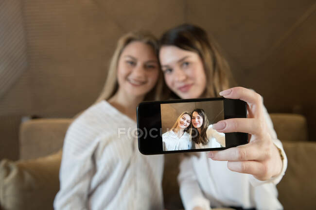 Duas amigas elegantes com xícaras de café fresco sorrindo e posando para selfie enquanto se senta à mesa no restaurante acolhedor — Fotografia de Stock
