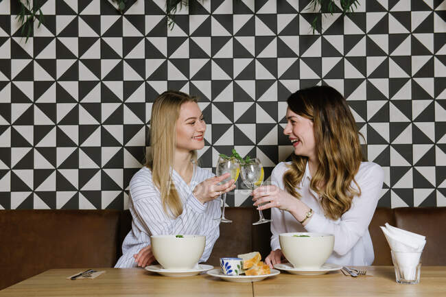 Dos hermosas damas sonriendo y tintineando vasos de cócteles de alcohol mientras almorzaban juntos en un acogedor restaurante - foto de stock