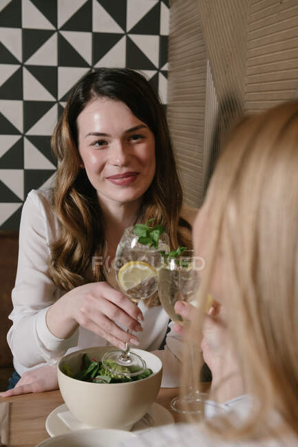 Two pretty ladies smiling and clinking glasses of alcohol cocktails while having lunch in cozy restaurant together — Stock Photo