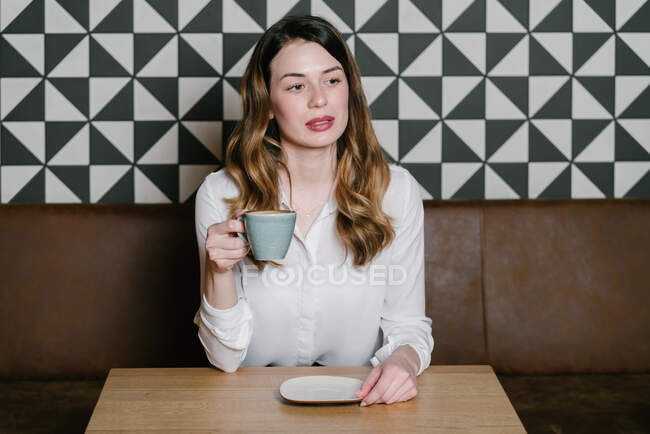 Pensive Жінка з чашкою свіжої кави, дивлячись далеко, сидячи за столом у затишному ресторані — стокове фото