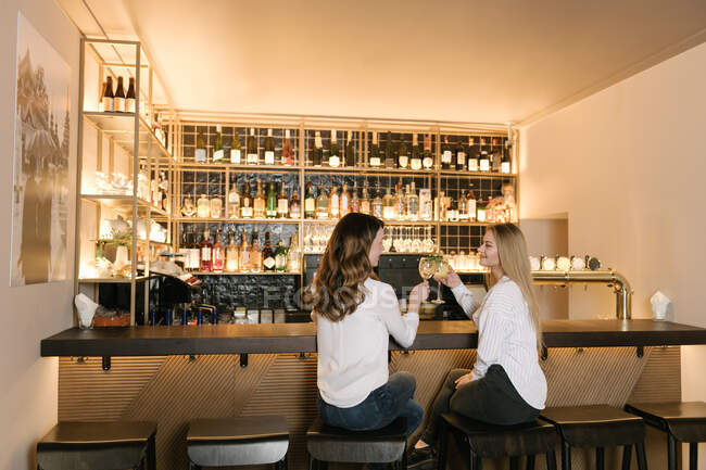 Обзор двух подруг, улыбающихся и звенящих стаканов алкогольных коктейлей во время пребывания в баре уютного ресторана — стоковое фото