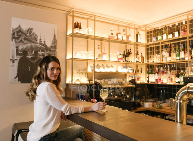 Вид збоку елегантна жінка зі склянкою алкогольного коктейлю дивиться на камеру, сидячи за барною стійкою в затишному ресторані — стокове фото