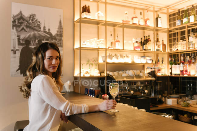 Вид збоку елегантна жінка зі склянкою алкогольного коктейлю дивиться на камеру, сидячи за барною стійкою в затишному ресторані — стокове фото