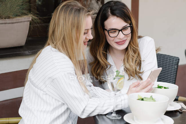 Elegant women friends using phone while sitting at table of outdoor restaurant during lunch — Stock Photo