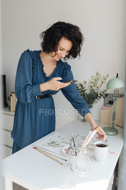 Allegro artista femminile in abito blu scattare foto sul telefono cellulare di lavoro acquerello sul tavolo in studio — Foto stock