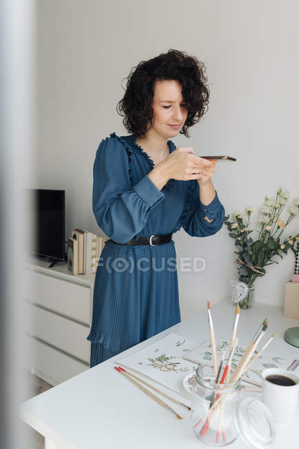 Donna in abito blu scattare foto sul telefono cellulare di lavoro acquerello sul tavolo in studio — Foto stock
