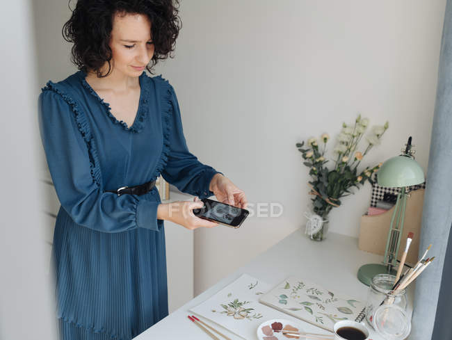Жінка-художник в блакитній сукні знімає на мобільному телефоні акварельні роботи на столі в студії — стокове фото
