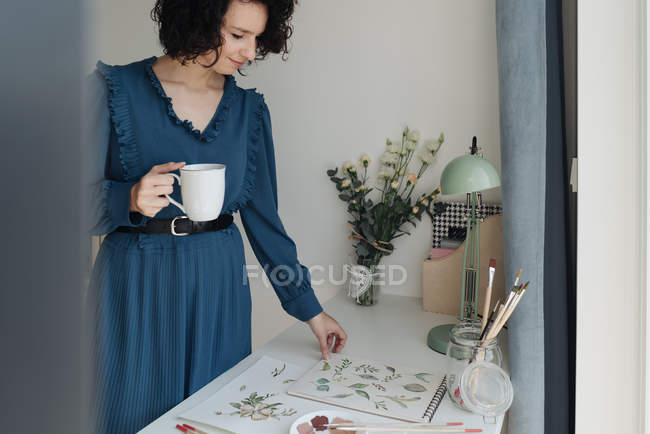 Brunette female artist standing next to a table with cup of coffee and looking at drawing in workplace — Stock Photo