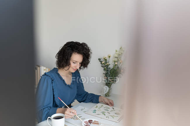 Элегантная женщина с кистью рисовать акварельные цветы на листе за столом — стоковое фото