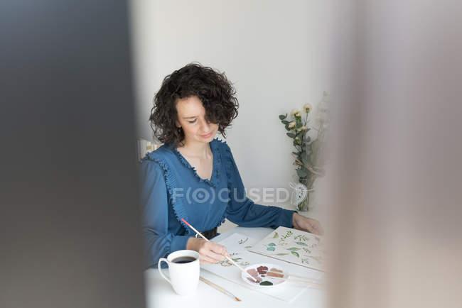 Mulher elegante com pincel pintando flores aquarela na folha na mesa — Fotografia de Stock