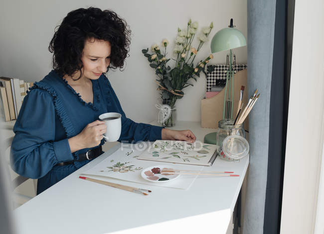 Брюнетка художница сидит за столом с чашкой кофе и смотрит на рисунок на рабочем месте — стоковое фото