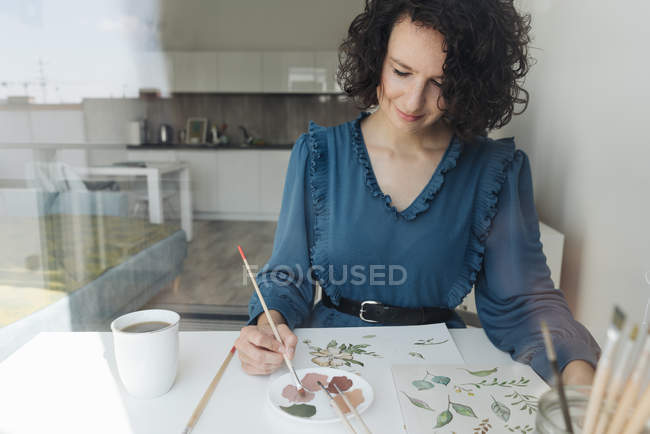 View through a window of elegant woman with brush painting watercolor flowers on sheet at desk — Stock Photo