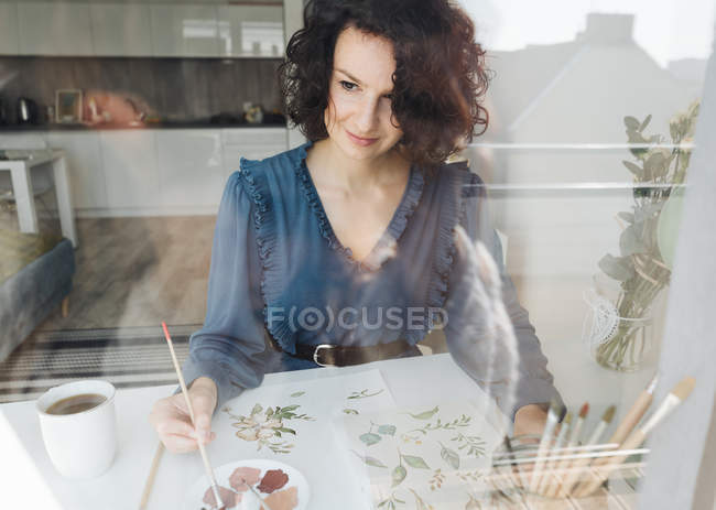 Vista attraverso una finestra di donna elegante con pennello pittura ad acquerello fiori su foglio alla scrivania — Foto stock