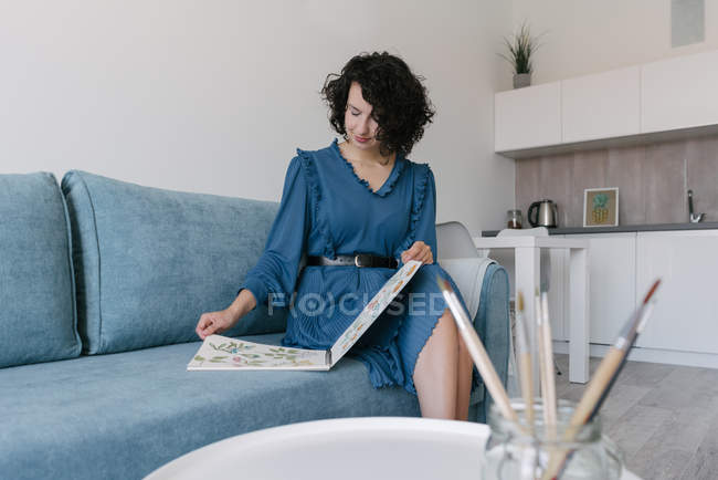 Bella giovane bruna seduta sul divano e alla ricerca di album con disegni a casa — Foto stock