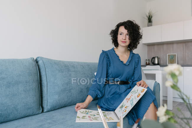 Pensativo hermosa joven morena hembra sentada en el sofá y mirando hacia otro lado sosteniendo un álbum con dibujos en casa - foto de stock