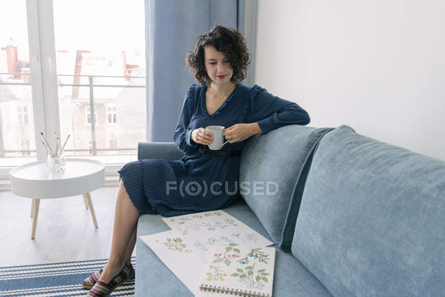 Молодая брюнетка сидит на диване и смотрит альбом с рисунками дома, наслаждаясь чашкой кофе — стоковое фото
