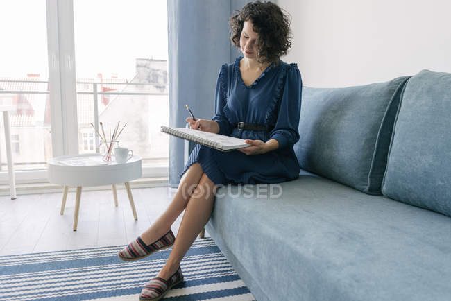 Elegante donna seduta su un divano che disegna su un taccuino a casa — Foto stock