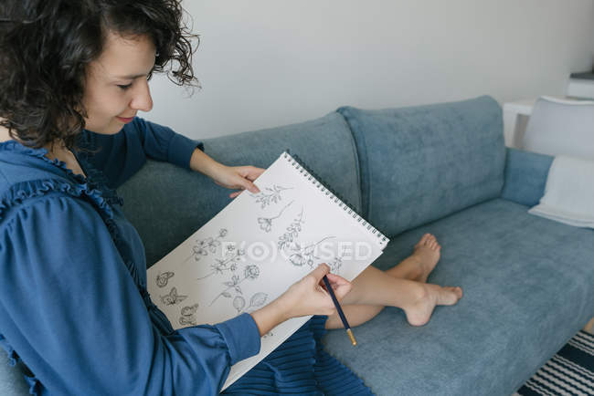 Elegant woman sitting on a sofa drawing flowers on a notebook at home — Stock Photo