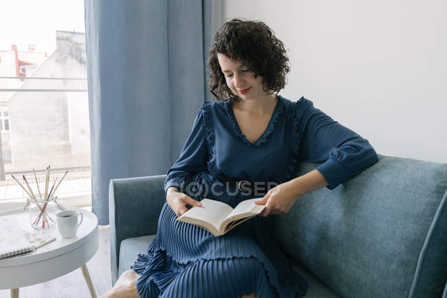 Happy young brunette woman in blue dress reading book sitting on couch at table with creative accessories tassels and album at home — Stock Photo