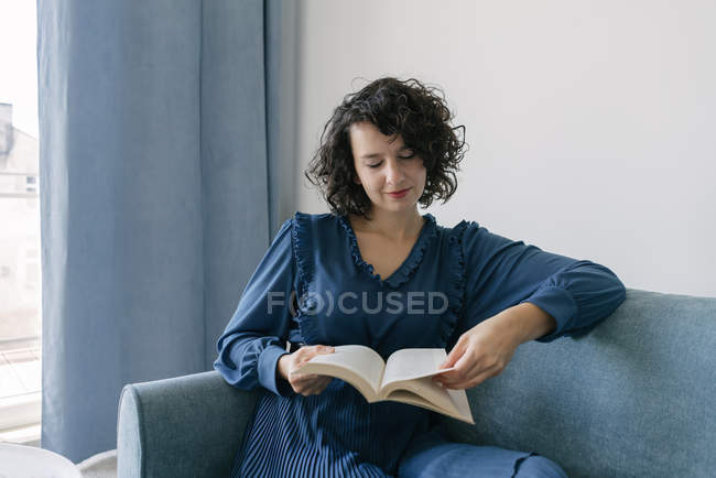 Happy young brunette woman in blue dress reading book sitting on couch at home — Stock Photo