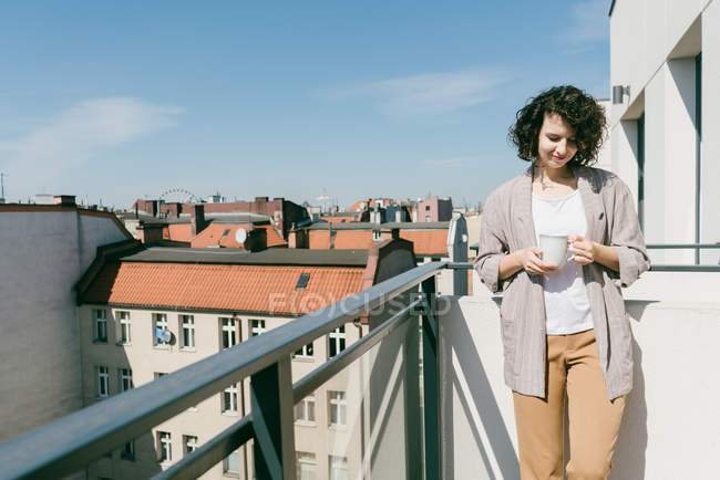 Mujer elegante disfrutando de la luz del sol en la terraza con los ojos cerrados mientras sostiene una taza de café - foto de stock