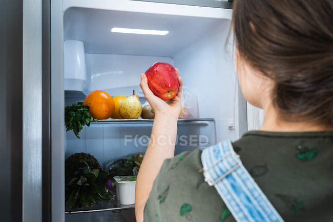 La hembra irreconocible tomando la manzana fresca de la balda del refrigerador en casa - foto de stock