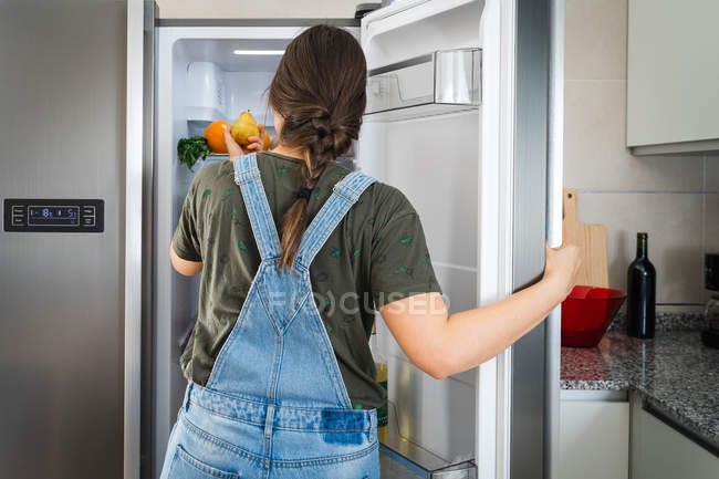 La hembra irreconocible tomando la pera fresca de la balda del refrigerador en casa - foto de stock
