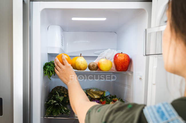 Неузнаваемая женщина берет свежий апельсин с полки холодильника дома — стоковое фото