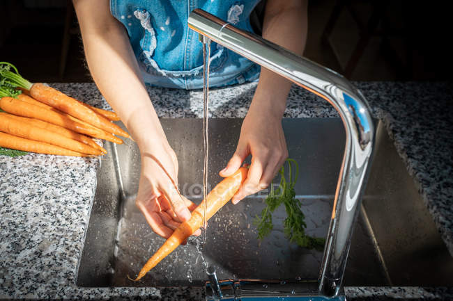 Imagen recortada de la mujer lavando zanahoria madura bajo agua limpia sobre el fregadero en casa - foto de stock